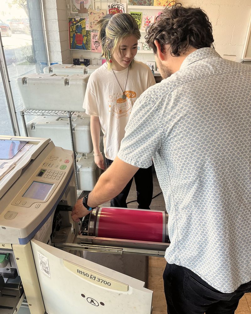 Two printers swapping the ink drum on a risograph machine