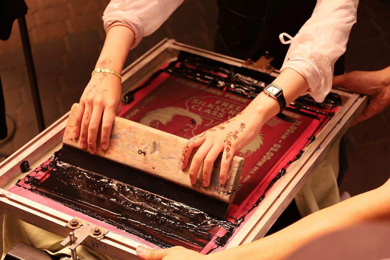 Two pairs of hands pulling ink across a screen on a printing bed