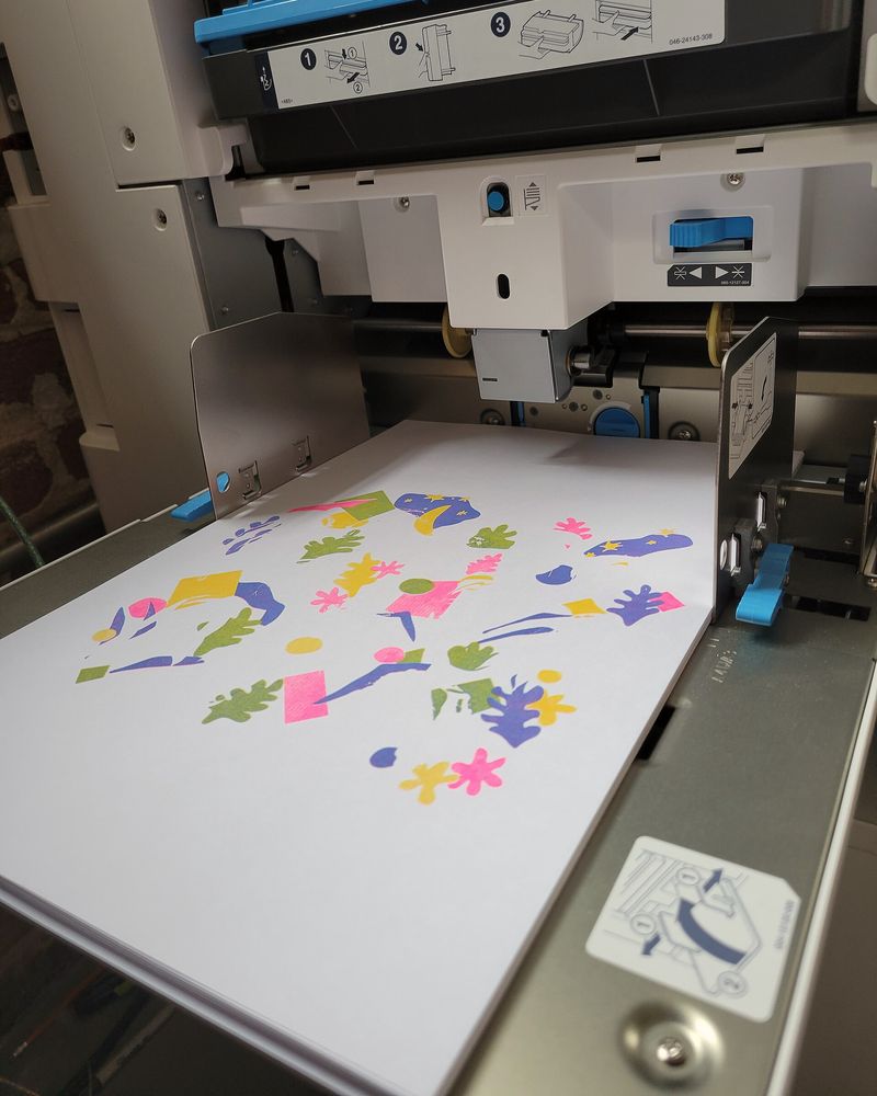 A sheet of pink, blue and yellow shapes coming out of a risograph onto the tray