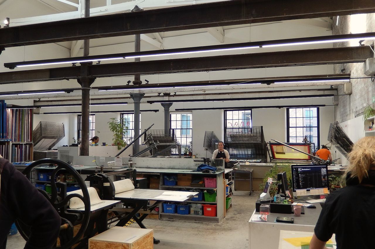 A large shared print studio with presses, tables and racks under north-light windows