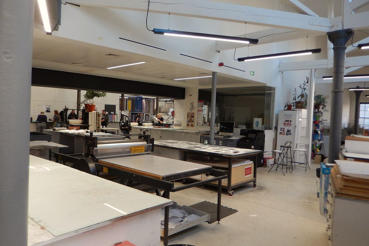 An open print workshop with long benches and a printer at work under roof lights