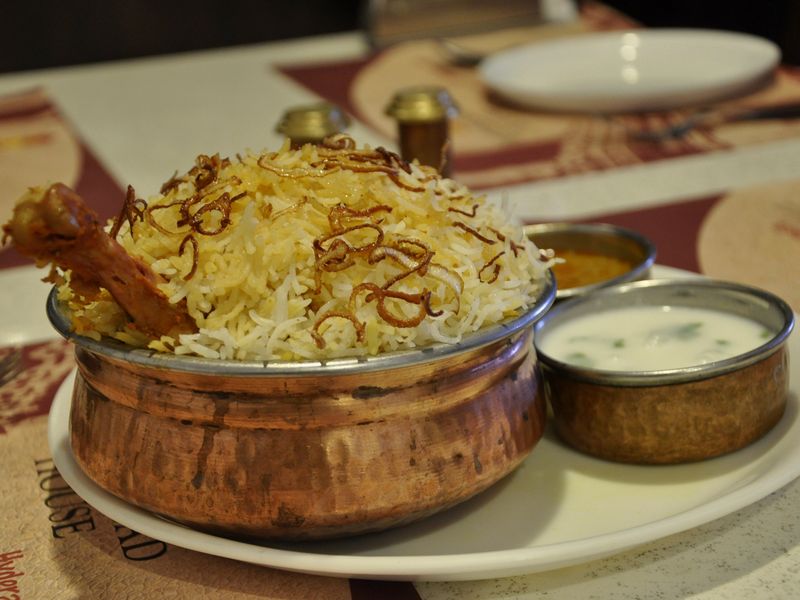 Chicken Hyderabadi biryani in a copper handi, with a bowl of raita beside it