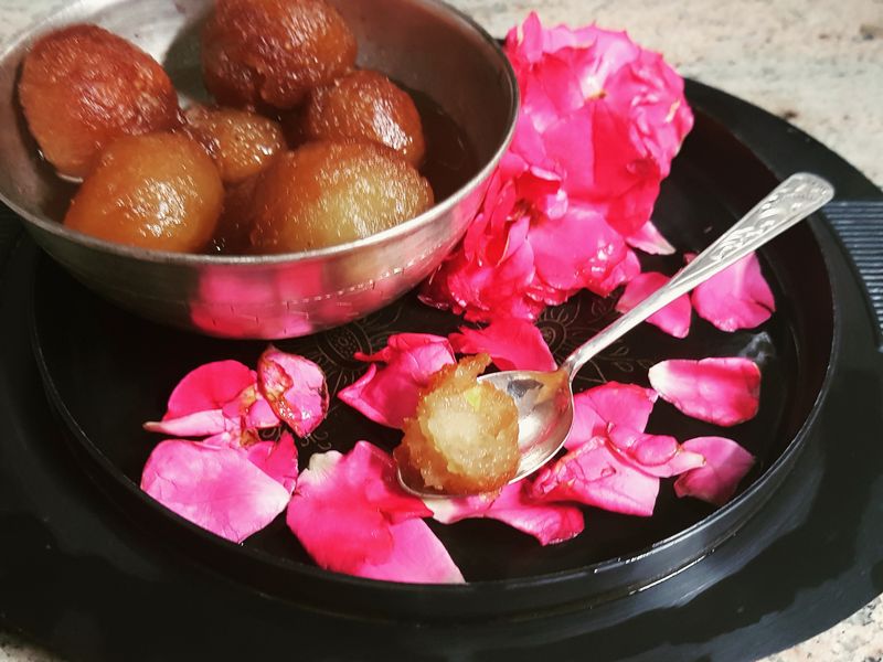 Gulab jamun in a steel bowl beside rose petals on a dark plate
