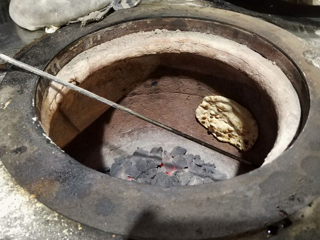The mouth of a clay tandoor with a flatbread baking on the wall over hot coals