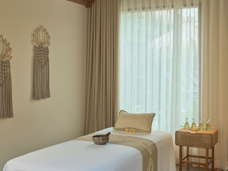 A calm treatment room with a made-up massage table, sheer curtains, a timber ceiling and a small tray of oils