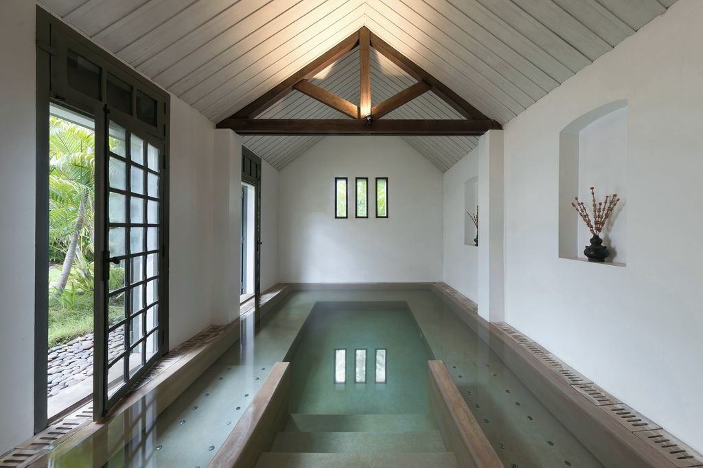 A long white room with a timber-framed ceiling, a pale plunge pool and tall windows onto a garden