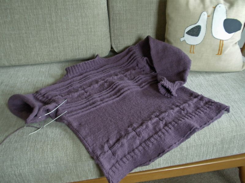 A purple hand-knitted gansey jumper in progress, laid on a sofa