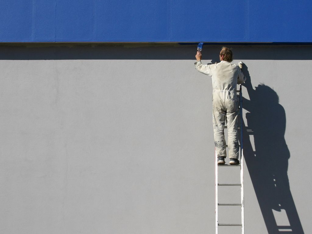 A decorator on a ladder painting a large white wall below a strip of blue