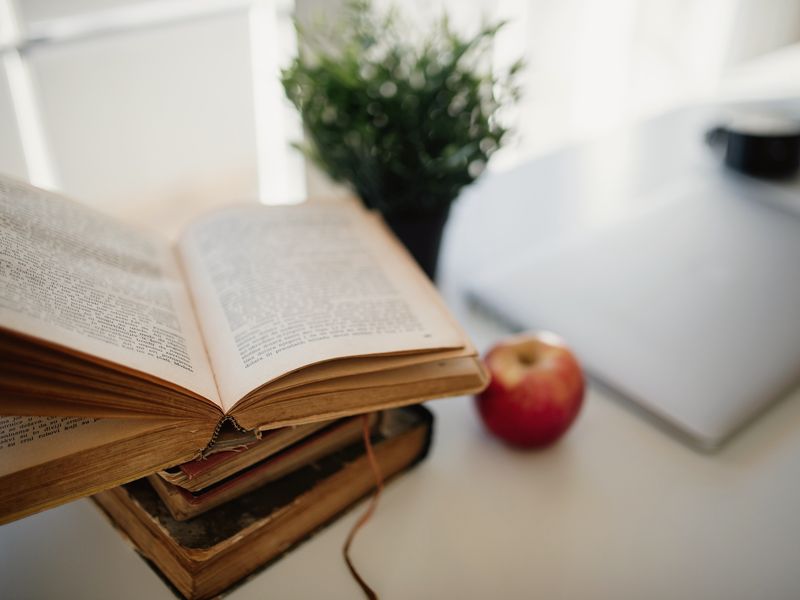 An open old book and a red apple on a bright desk beside a laptop