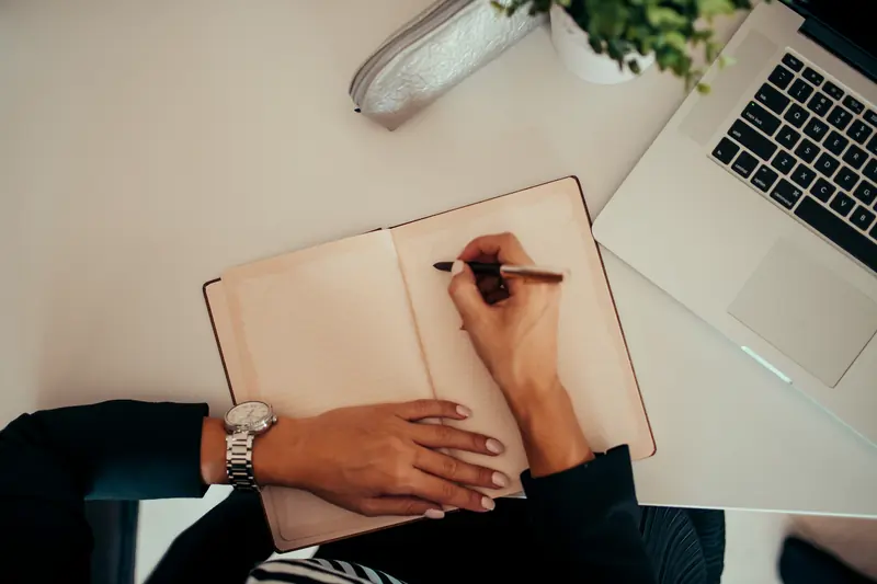 Hands writing in an open notebook at a desk beside a laptop