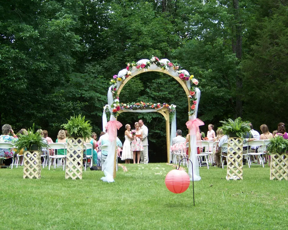 Garden wedding arch decorated with white and pink flowers on a lawn, with rows of chairs and guests among the trees behind