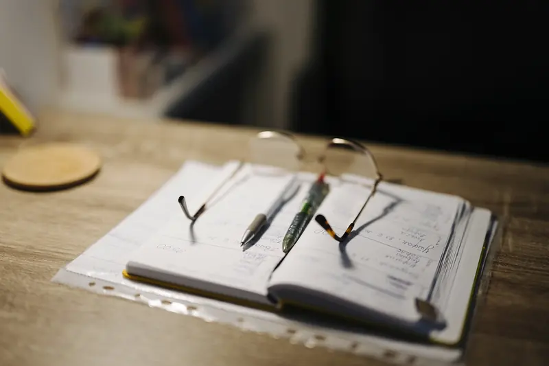An open notebook with pens and reading glasses on a desk in lamplight