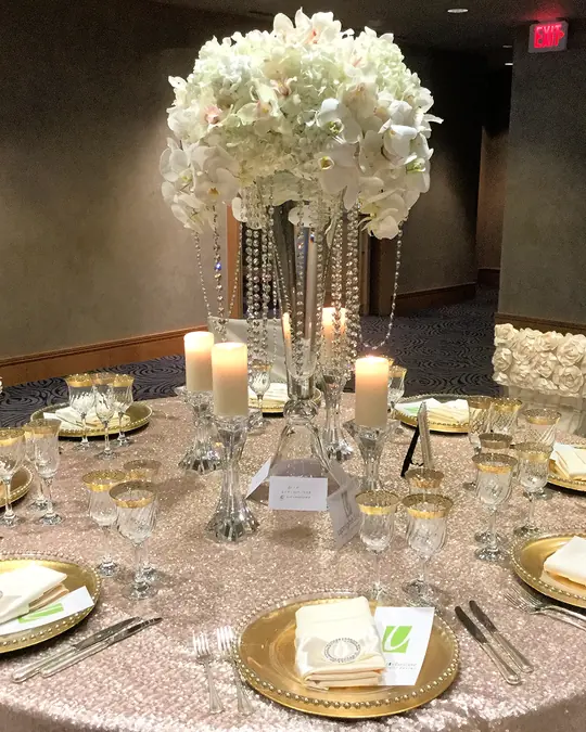 A round table set with gold chargers and glassware under a tall arrangement of white flowers