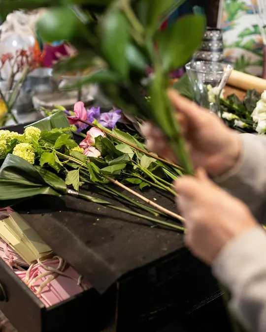 A florist's hands arranging a bouquet of greenery and pink flowers at a workbench