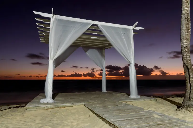 A white wooden pavilion hung with sheer drapes on a beach deck at dusk