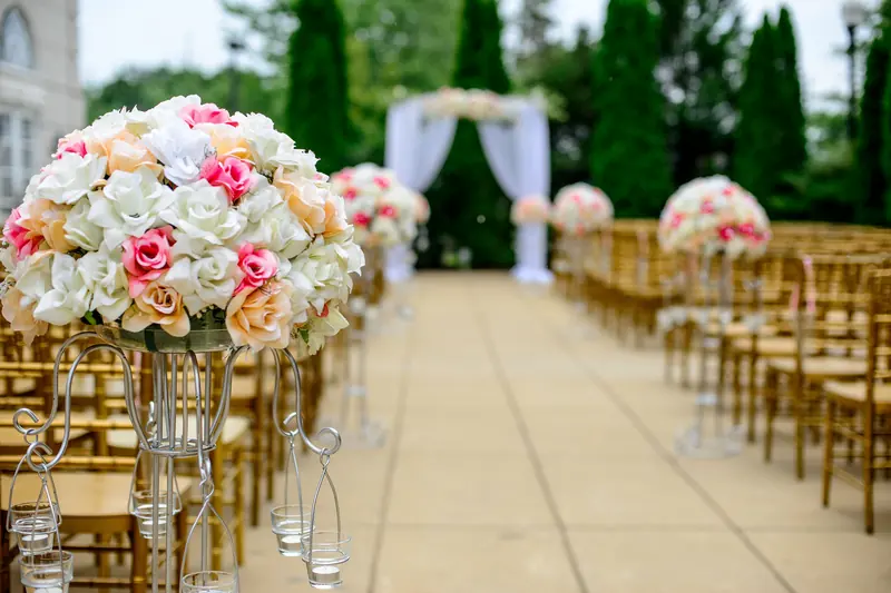 Outdoor wedding aisle lined with wooden chairs and tall arrangements of pink and white roses, leading to a white draped arch