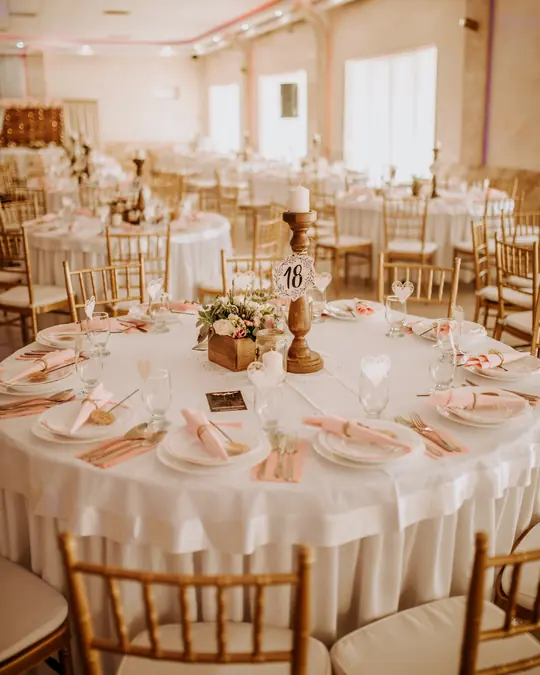Round tables with white cloths, gold chairs and flowers in a warmly lit banquet hall